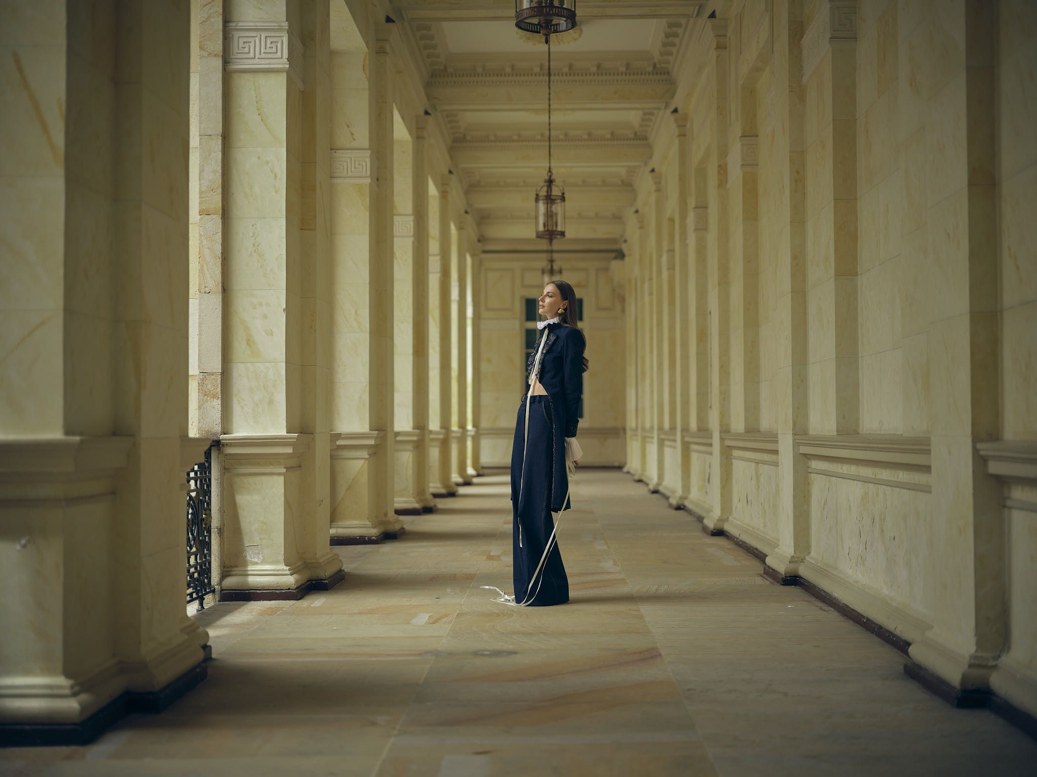 Modelo con pantalón negro de bota amplia y abrigo largo en un corredor de columnas, colección Guadalupe de Diego Guarnizo