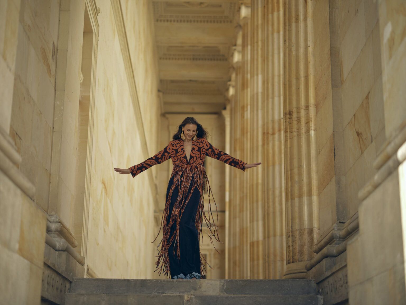 Modelo con chaqueta de flecos y los brazos abiertos en un corredor de columnas, colección Guadalupe de Diego Guarnizo